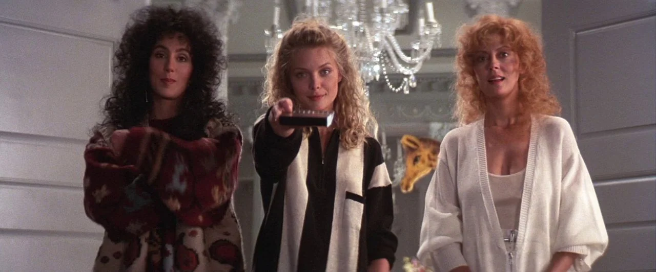 The Witches of Eastwick (1987) movie still: medium — Three women stand in a grand, brightly lit interior space with a hanging crystal chandelier.…