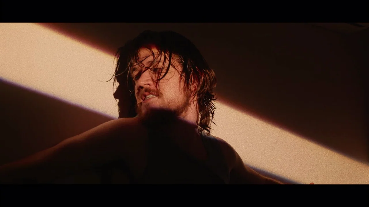 Bo Burnham: Inside (2021) movie still: closeup — A dark room features a single sharp sliver of light illuminating a man's face. The man with damp,…