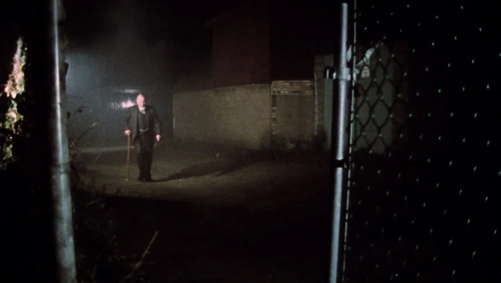Martin (1977) movie still: wide — A dark, foggy urban alleyway at night illuminated by a single overhead light. An elderly man in a suit walks…