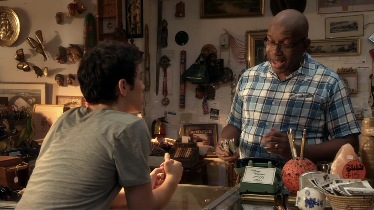 Rags (2012) movie still: medium — An interior space is crowded with various vintage objects and wall-mounted art. A man in a blue shirt gestures…