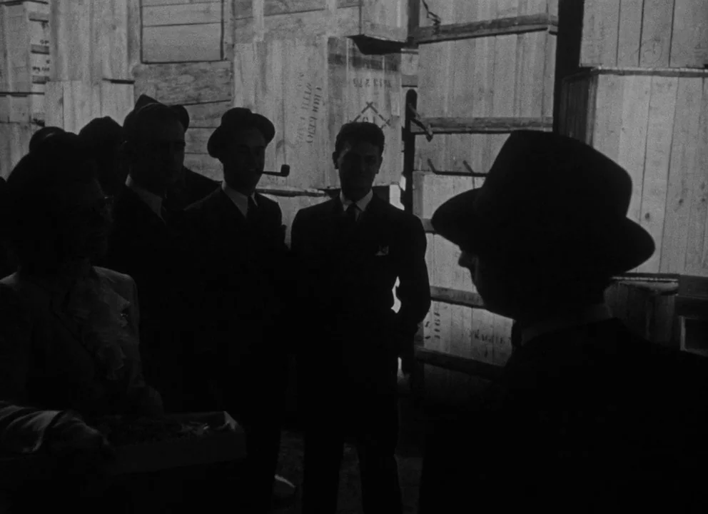 Citizen Kane (1941) movie still: medium — A dark, cramped space filled with stacked wooden shipping crates and packing materials. A small group of…