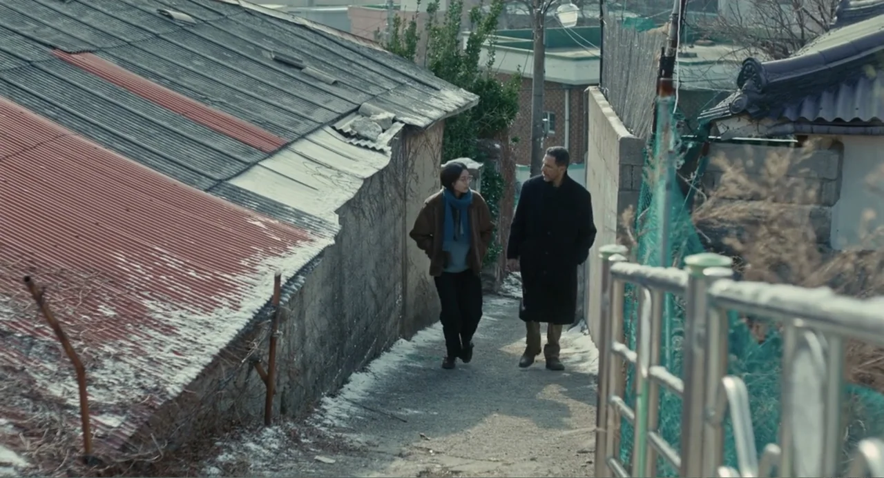 Winter in Sokcho (2025) movie still: fullbody — A narrow alleyway sits between a corrugated metal roof and a high concrete wall. A woman and a man…