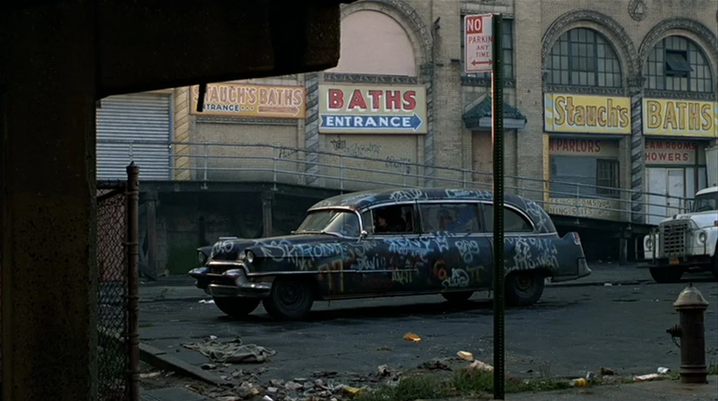 The Warriors (1979) movie still: wide — An aged brick building with arched windows features faded signs for Stauch's Baths. A black vintage car…