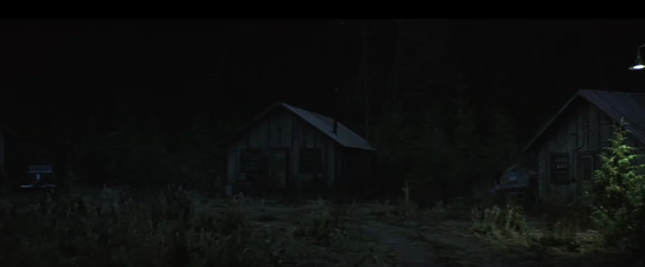 This Boy's Life (1993) movie still: wide — Dark wooden cabins sit nestled in a dense, unlit forest at night. Two older automobiles are parked…