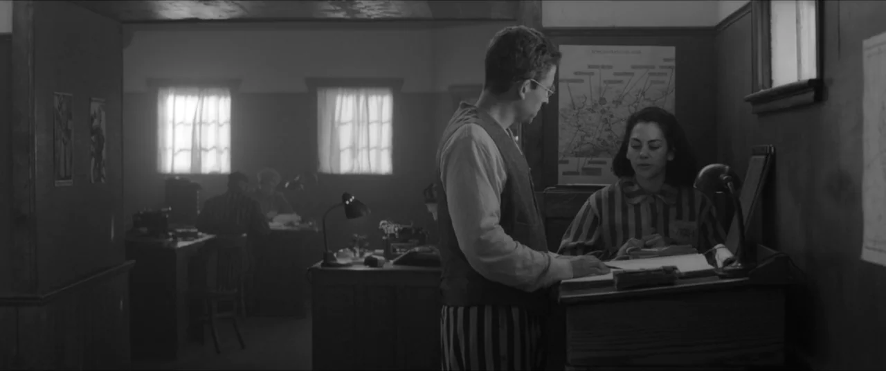 Bau, Artist at War (2025) movie still: medium — A sparse administrative room features dark wooden walls with a map hanging on one side. A man and a…