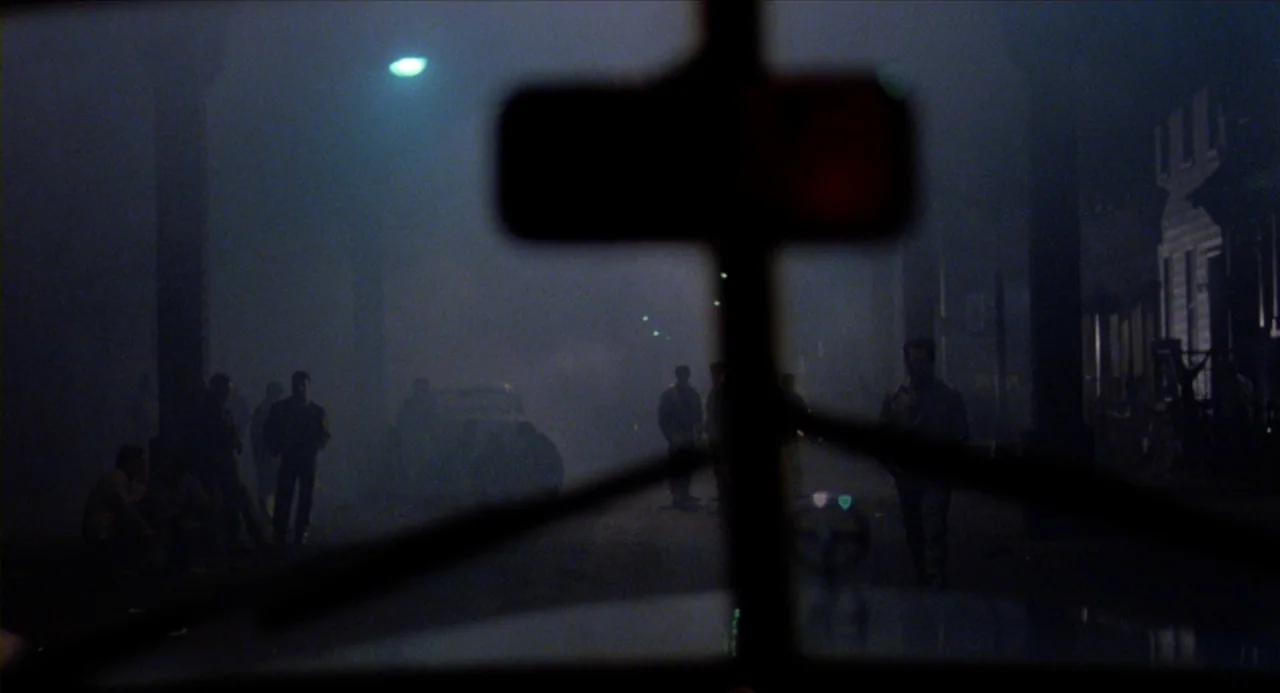 The Wanderers (1979) movie still: wide — A dark city street is obscured by dense fog at night under a streetlamp. Silhouettes of men stand on the…