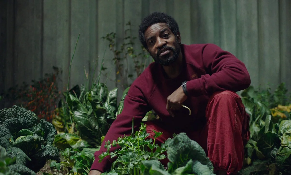 High Life (2018) movie still: medium — A man crouches in a dense vegetable garden next to a vertical ribbed metal wall. He tends to the low-growing…