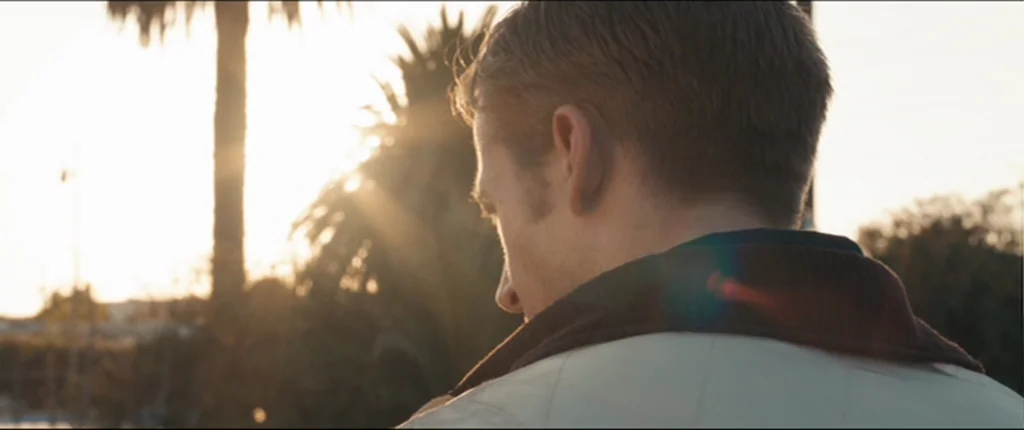 Drive (2011) movie still: closeup — A man stands outdoors with palm trees visible in the soft background sunlight. The man looks toward the horizon…