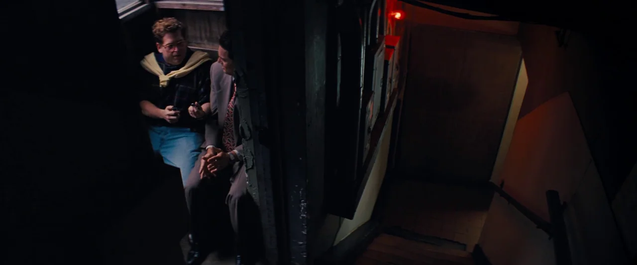 The Wolf of Wall Street (2013) movie still: medium — A narrow stairwell entrance with dark walls and a singular glowing red light source. Two men in…