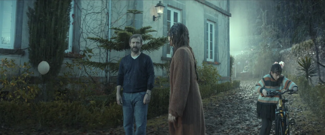 Tell Me Softly (2025) movie still: fullbody — A stone house exterior features a gravel path covered in fallen autumn leaves. A man and a woman stand…