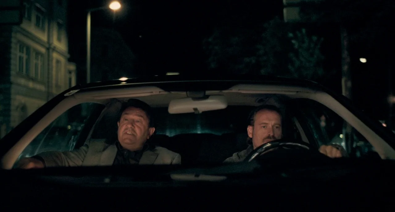Revanche (2008) movie still: medium — Two men sit inside the front seats of a moving car at night. The windshield captures a blurry view of a city…
