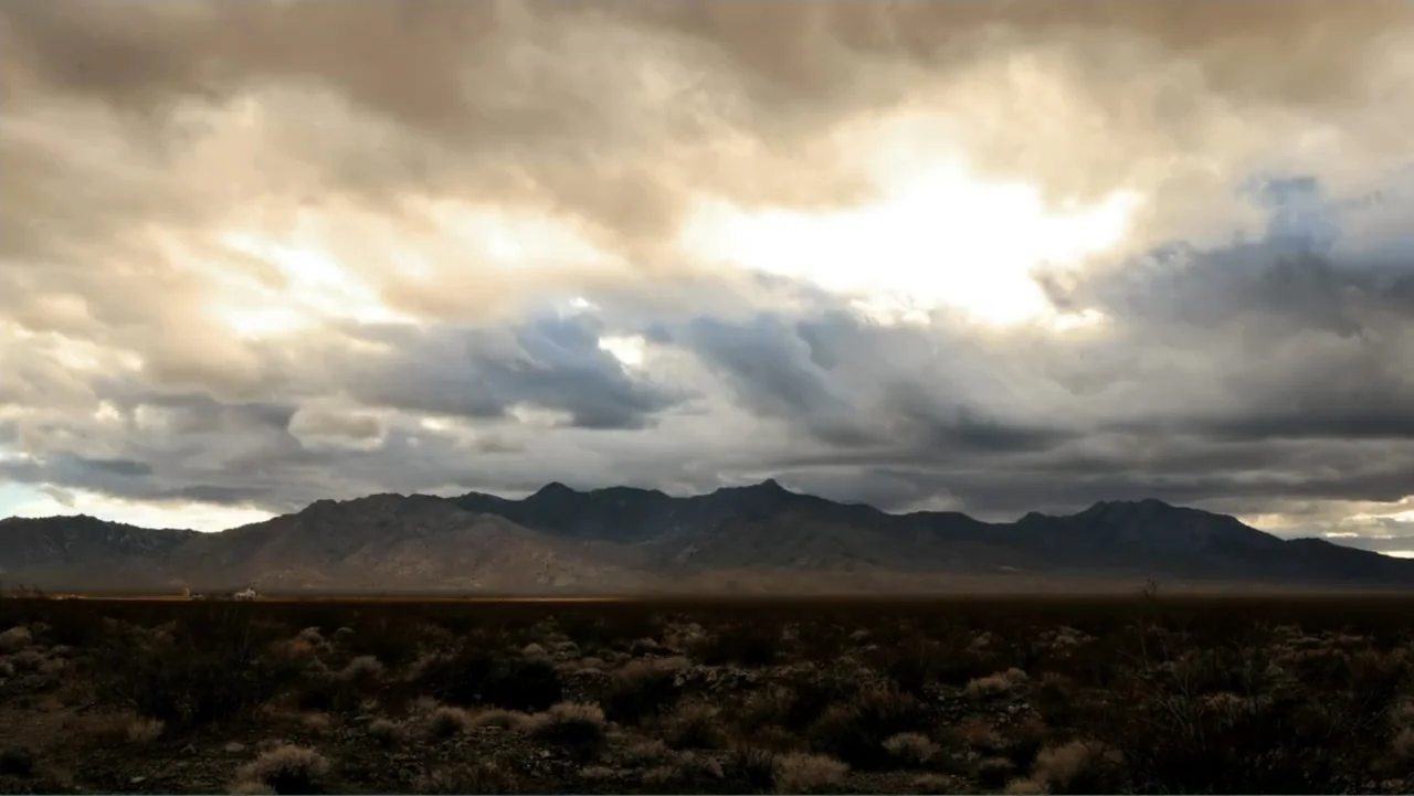 Savageland (2015) movie still: establishing — A vast, arid desert expanse stretches toward a range of dark mountains. Heavy, dramatic storm clouds…