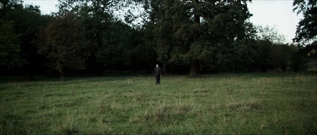 Kill List movie still: wide — A vast, grassy meadow bordered by a dense line of mature deciduous trees. A lone male figure stands in the center of…