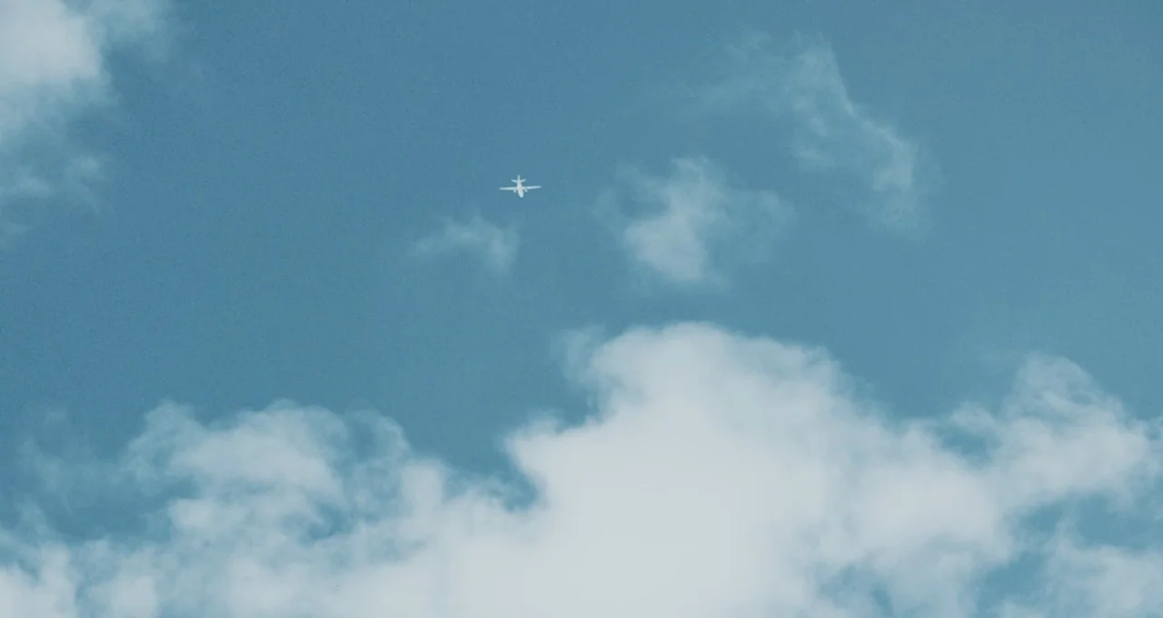 Balloon (2019) movie still: establishing — A vast blue sky is filled with scattered white cumulus clouds. A small passenger airplane flies at a high…
