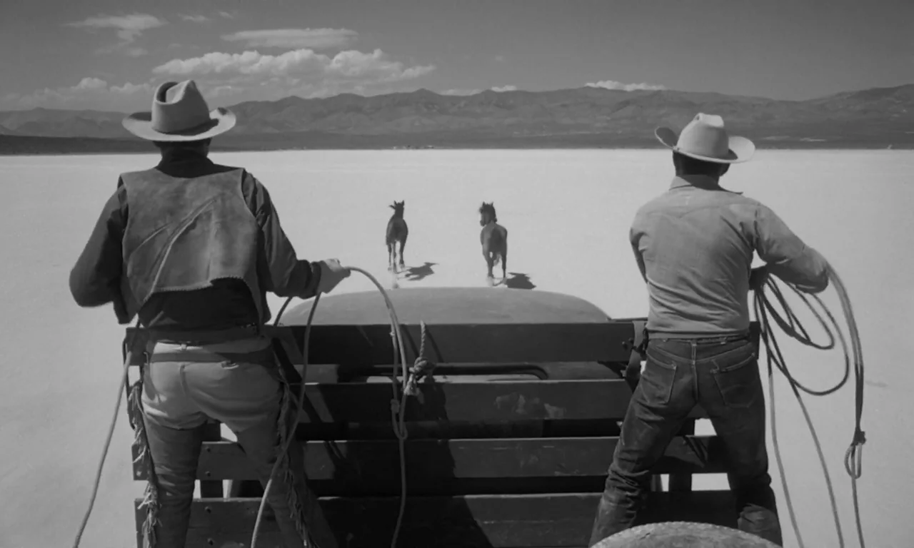 The Misfits (1961) movie still: fullbody — Two cowboys stand on the tailgate of a moving truck on a vast salt flat. They hold ropes extended toward…