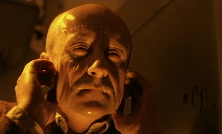 Golem (1980) movie still: closeup — A bald man is captured in a close frame against a plain wall. He presses his hands against his ears with his…