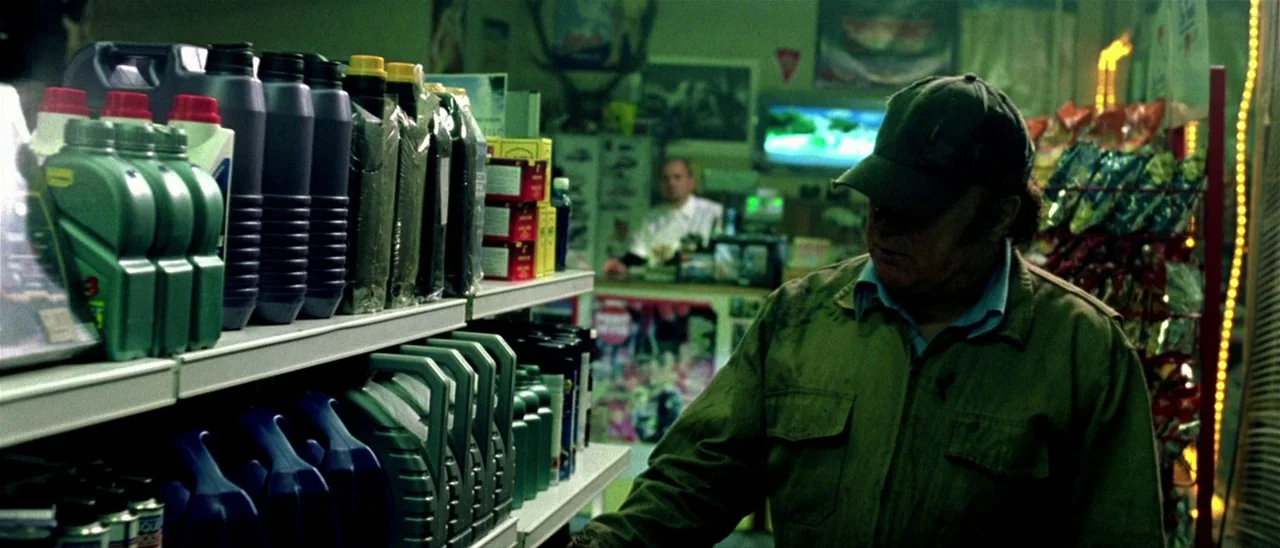 Haute Tension (2003) movie still: medium — Rows of plastic oil jugs fill the foreground of a dim retail interior. A man in a baseball cap looks at…
