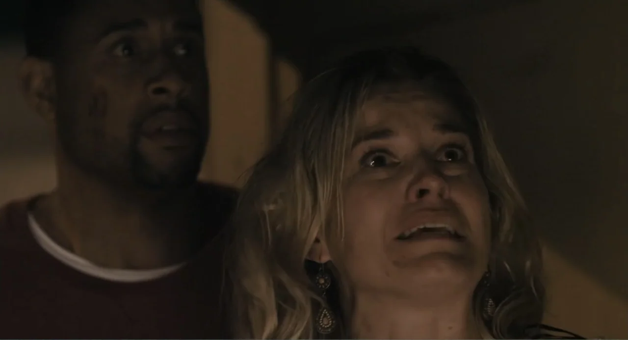 Gates of Darkness (2019) movie still: medium — An interior dimly lit corridor features two people looking forward in a darkened space. A man and a…