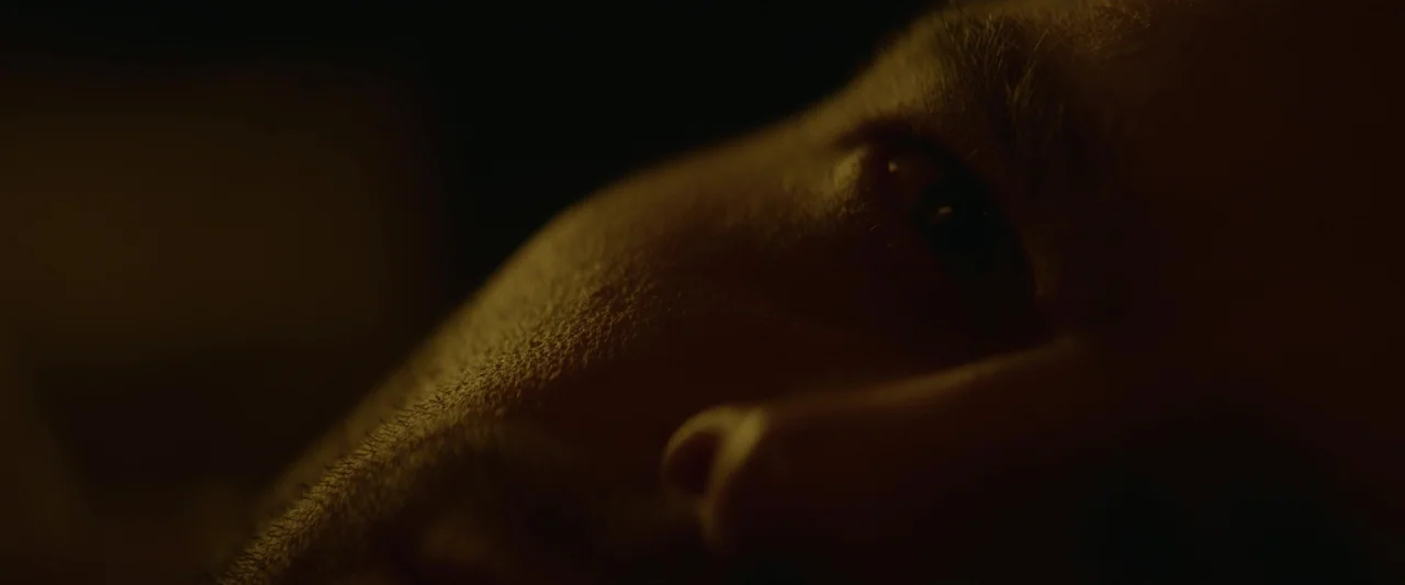 Fair Play (2023) movie still: closeup — A close view of a face lying on a pillow in deep darkness. The skin is illuminated by a narrow beam of amber…
