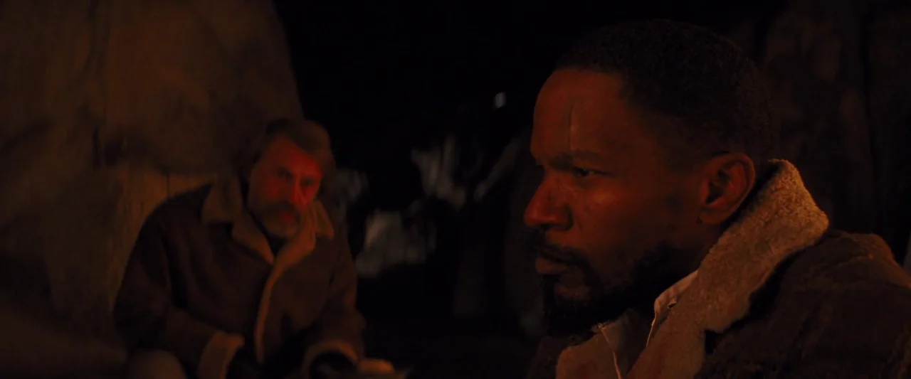 Django Unchained (2012) movie still: medium — A dark, rocky nighttime campsite lit only by the glowing light of a fire. Two men in heavy winter…