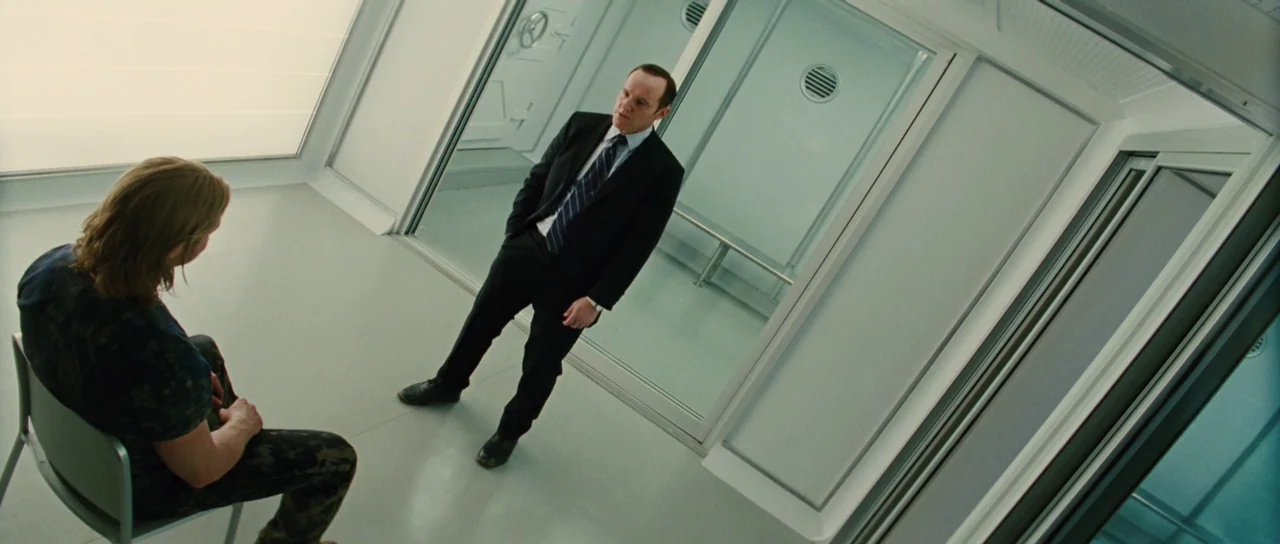 Thor (2011) movie still: wide — A sterile interrogation room features white walls, metal accents, and glass partitions. A man in a suit stands…