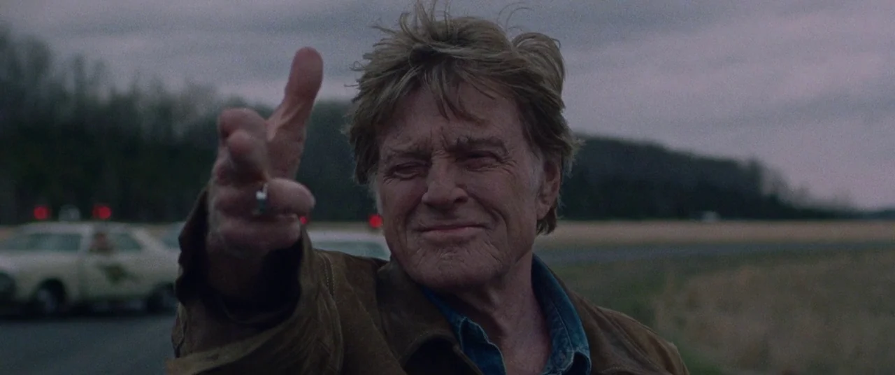 The Old Man & The Gun (2018) movie still: closeup — An overcast field with a blurred car in the background under gray clouds. An older man points…