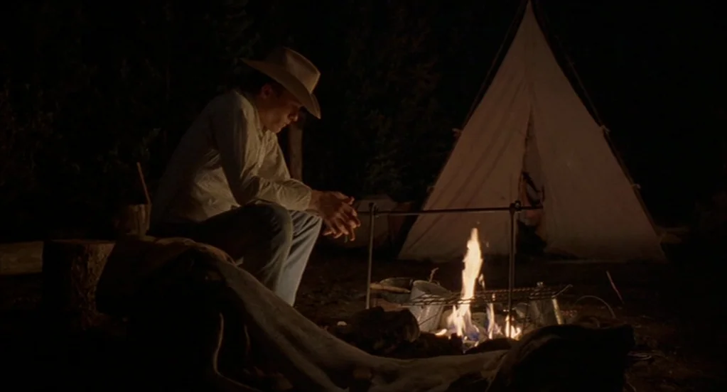 Brokeback Mountain (2005) movie still: medium — A dark, wooded campsite at night features a small white tent in the background. A young man in a…