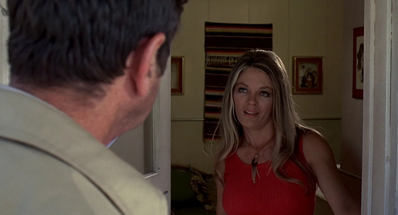 Charley Varrick (1973) movie still: medium — A woman stands in a doorway looking at a man in a tan jacket. The interior walls feature a striped wall…