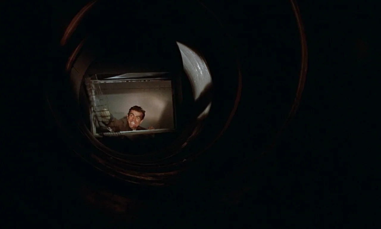Dr. No (1962) movie still: medium — A dark, circular ventilation pipe frames a brightly lit rectangular opening showing a man. The man grips the…