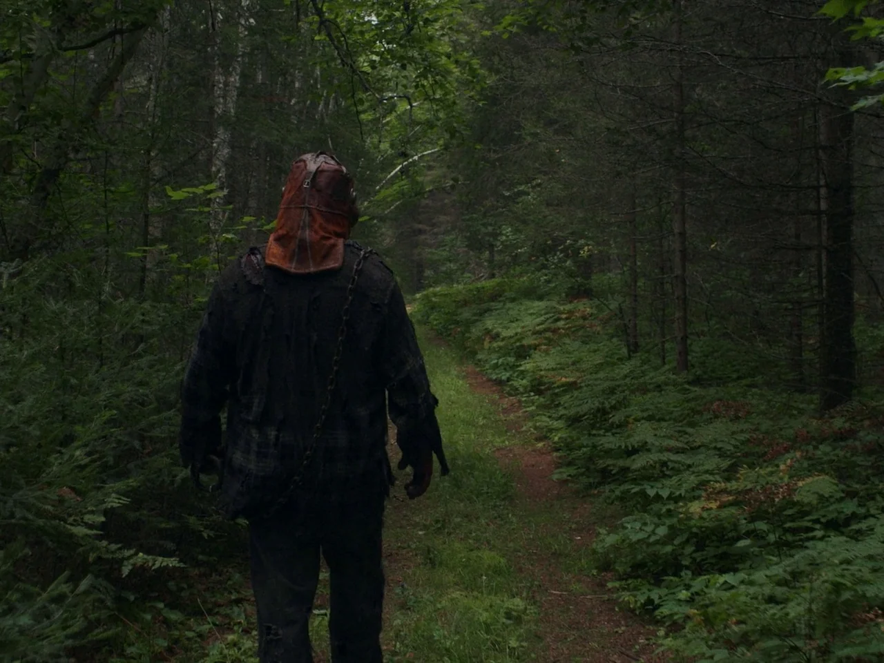 In A Violent Nature (2024) movie still: fullbody — A dense forest path winds through thick green undergrowth and tall evergreen trees. A masked…