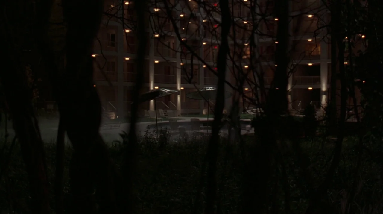 Lady in the Water (2006) movie still: establishing — A multi-story hotel courtyard at night features an empty pool area with umbrellas. Bare tree…