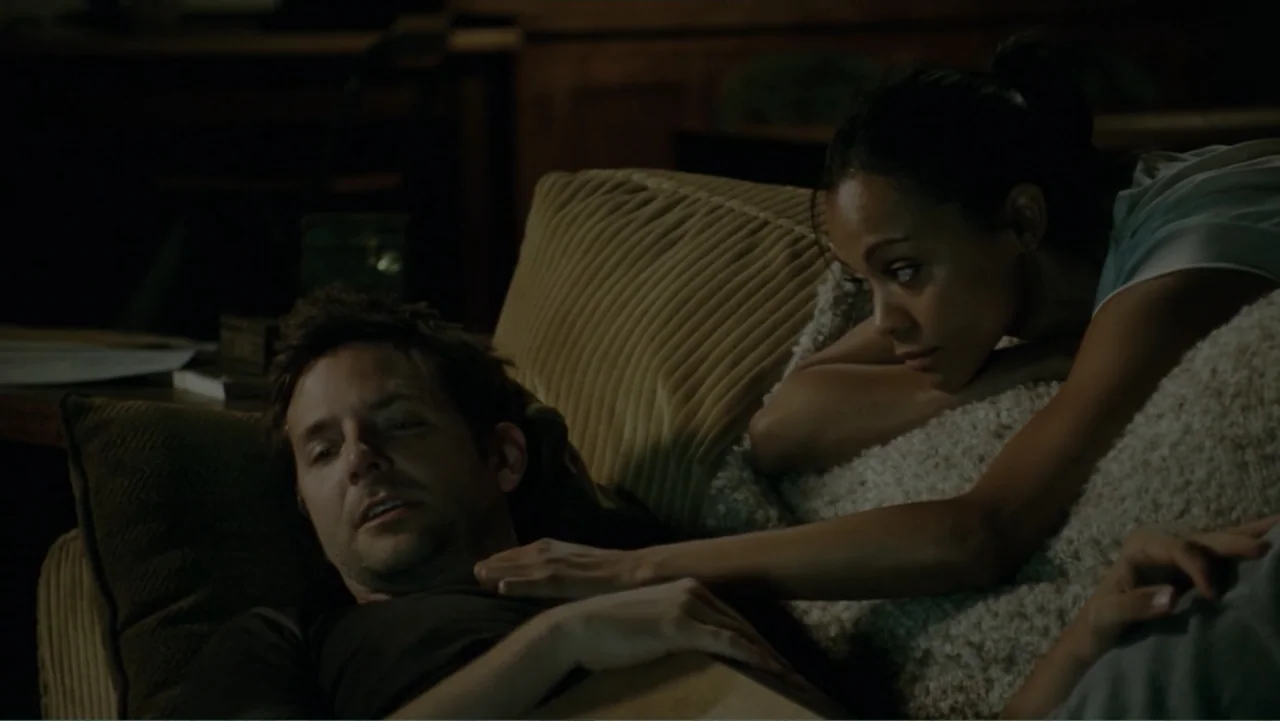 The Words (2012) movie still: medium — A dimly lit room contains a large upholstered sofa with textured cushions. A man lies back on the cushions…