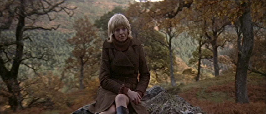 Images (1972) movie still: medium — A woman in a brown coat rests on a rock on a hillside. Bare trees and autumn foliage surround her in an open…