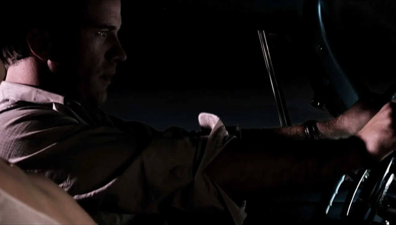 Dark Country (2009) movie still: medium — A dark car interior at night with a man focused on the road ahead. The subject is driving with his hands…