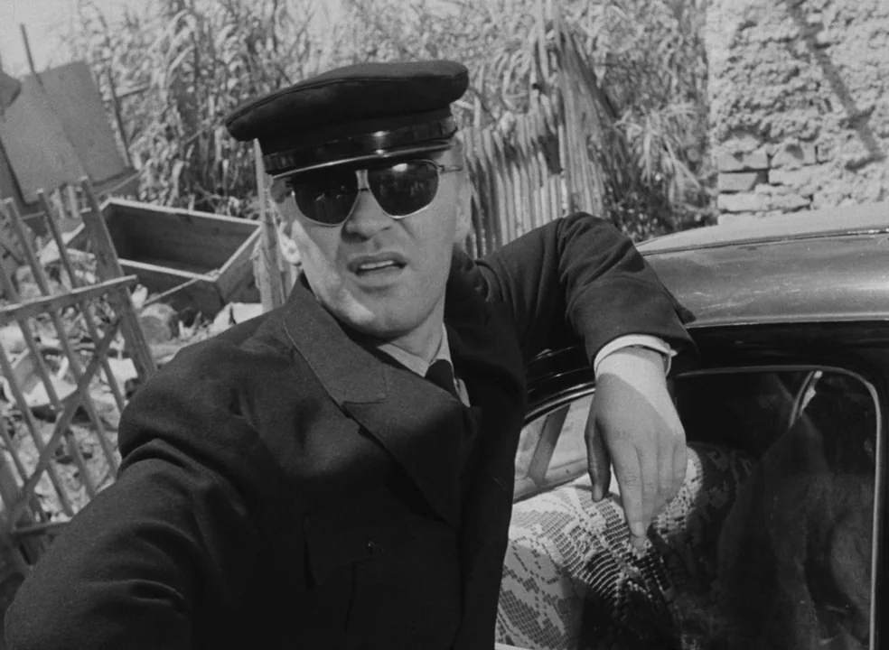 Il Bidone (1955) movie still: medium — A man in a uniform leans against a car door in an outdoor rural setting. Tall dry vegetation and a brick wall…