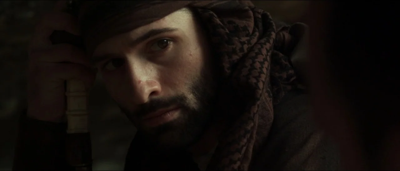 The Penitent Thief (2021) movie still: closeup — A dimly lit interior space with dark, indistinct surroundings. A man with a dark beard wearing a…