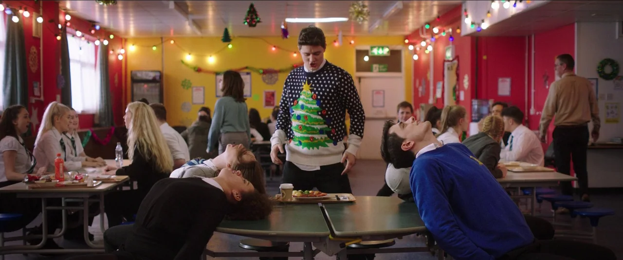 Anna and the Apocalypse (2018) movie still: medium — A school cafeteria decorated with Christmas tinsel and holiday string lights. Two students tilt…