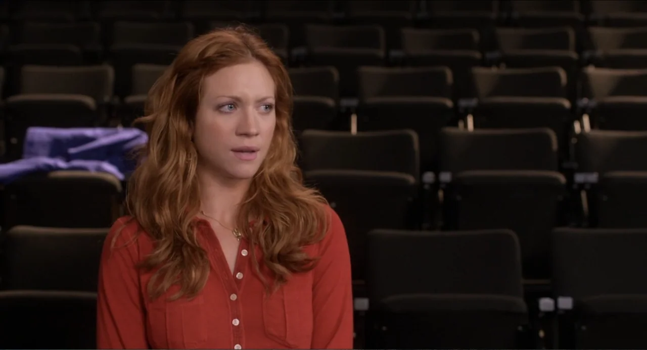 Pitch Perfect (2012) movie still: medium — A woman sits in the foreground of a darkened, empty theater auditorium. She wears a red shirt and gazes…