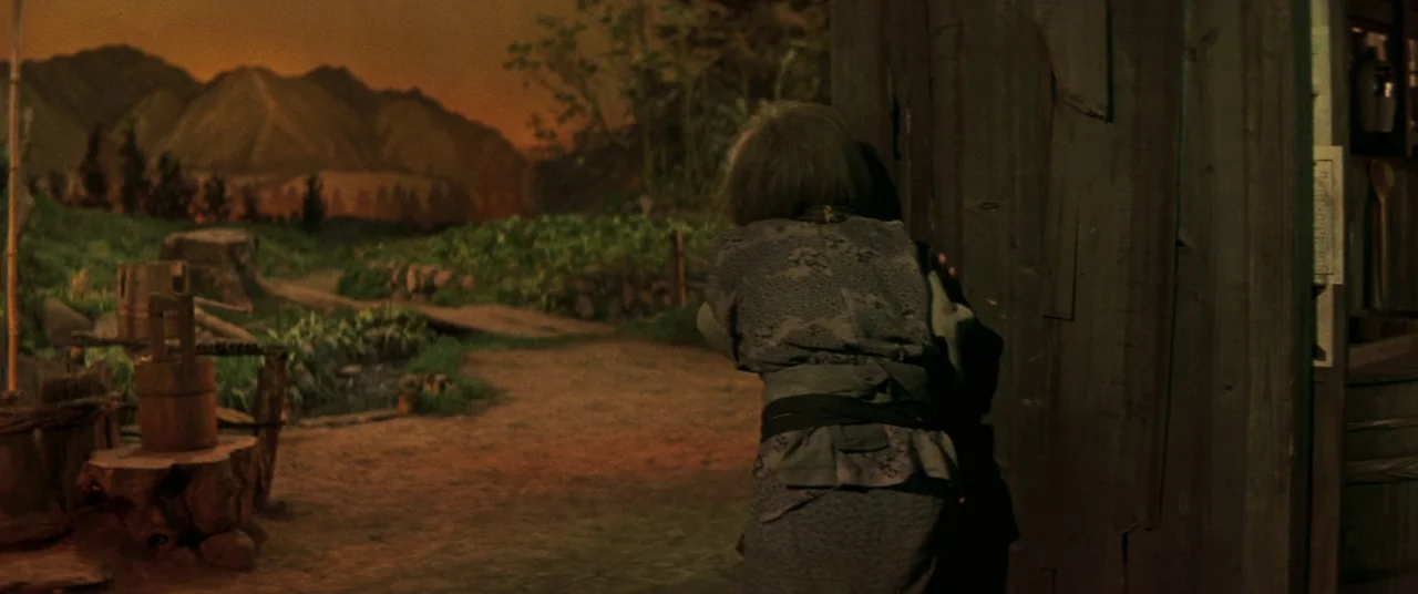 Ballad Of Narayama (1958) movie still: medium — A rural farmstead featuring a painted landscape backdrop of mountains at dusk. An elderly woman in a…
