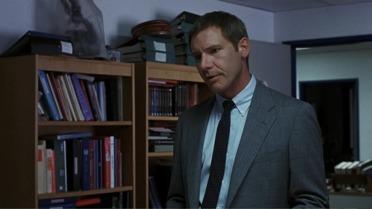 Presumed Innocent (1990) movie still: medium — An office interior with wooden bookshelves packed with binders and books. A man in a suit stands in…