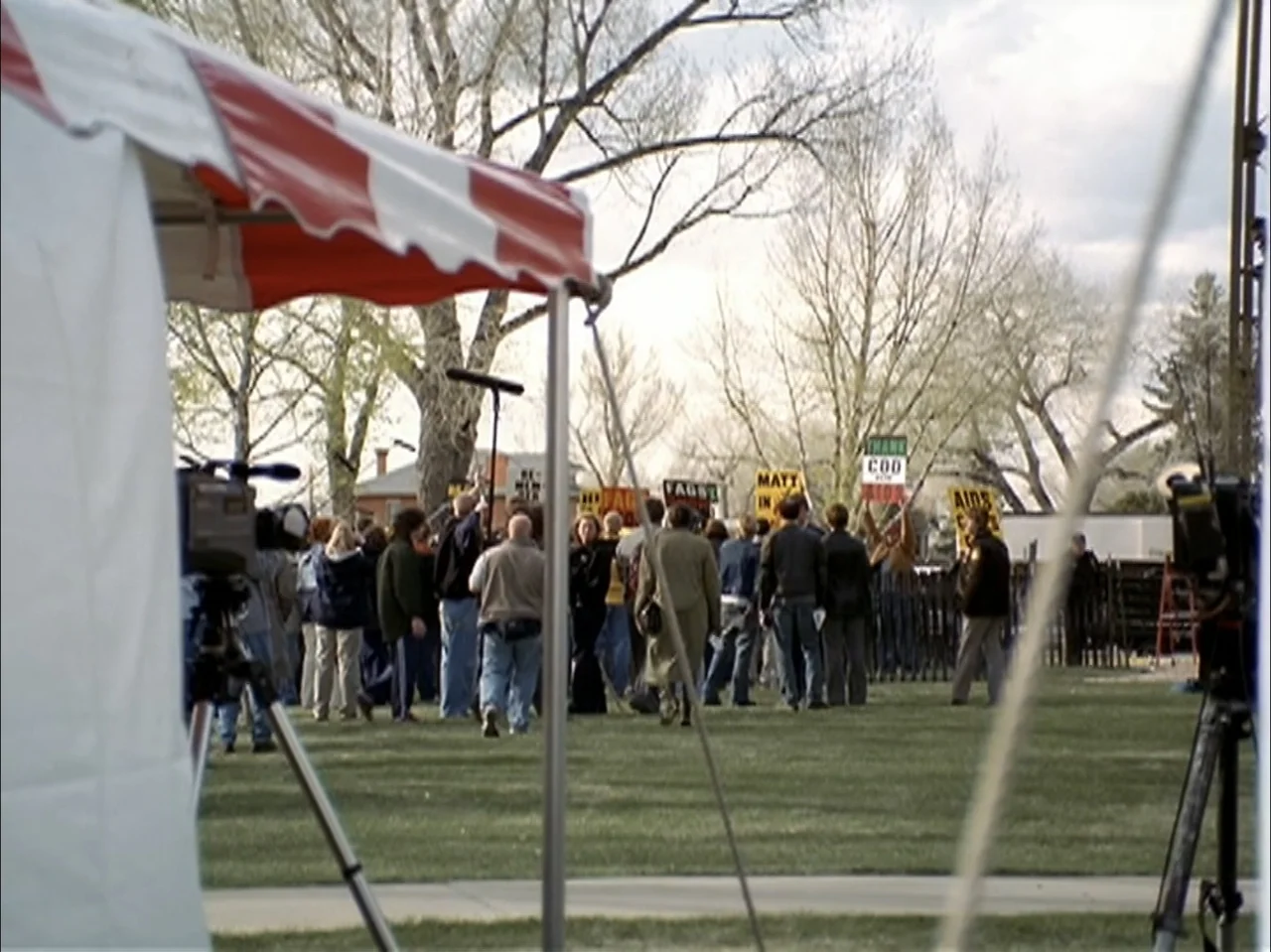 The Laramie Project (2020) movie still: wide — A public lawn features a crowd of people gathered with protest signs. Individuals stand on the grass…