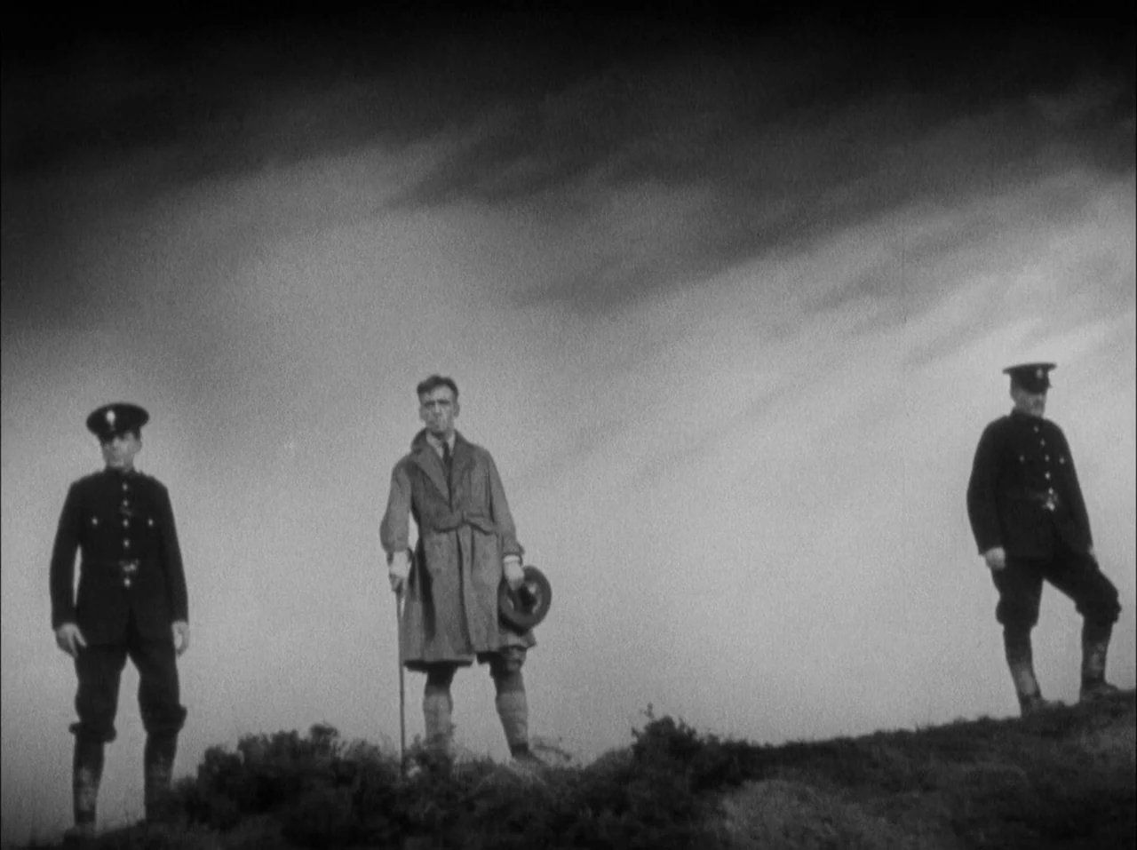 The 39 Steps (1935) movie still: fullbody — Three men stand on a grassy ridge against a dark and cloudy sky. The central figure holds a hat and cane…