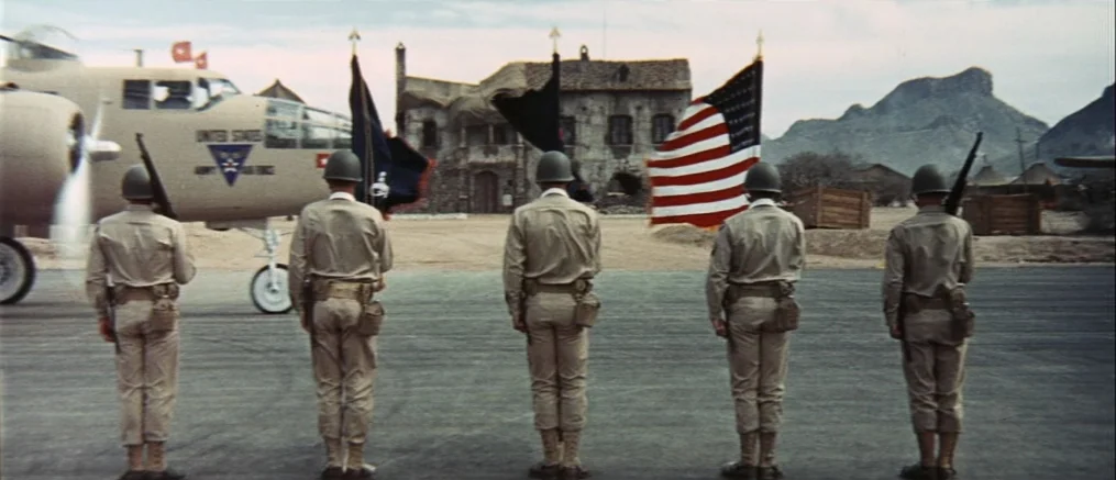 Catch 22 (1970) movie still: wide — A group of soldiers stands in formation on a paved airfield before a bomber plane. An American flag and other…