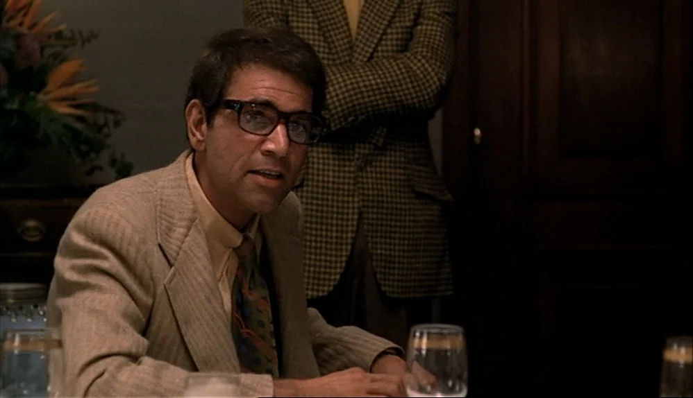 The Godfather (1972) movie still: medium — A dimly lit room features a table with wine glasses and floral arrangements. A man wearing glasses sits…