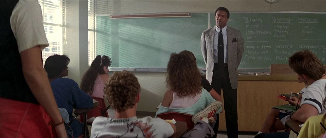 Bill and Ted’s Excellent Adventure (1989) movie still: medium — A high school classroom contains students sitting at desks in front of a chalkboard.…