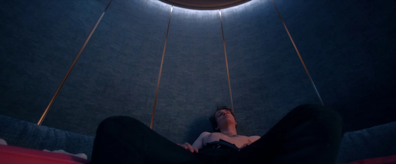 The Substance (2024) movie still: medium — A small circular pod features curved walls covered in blue fabric and copper accents. A shirtless young…