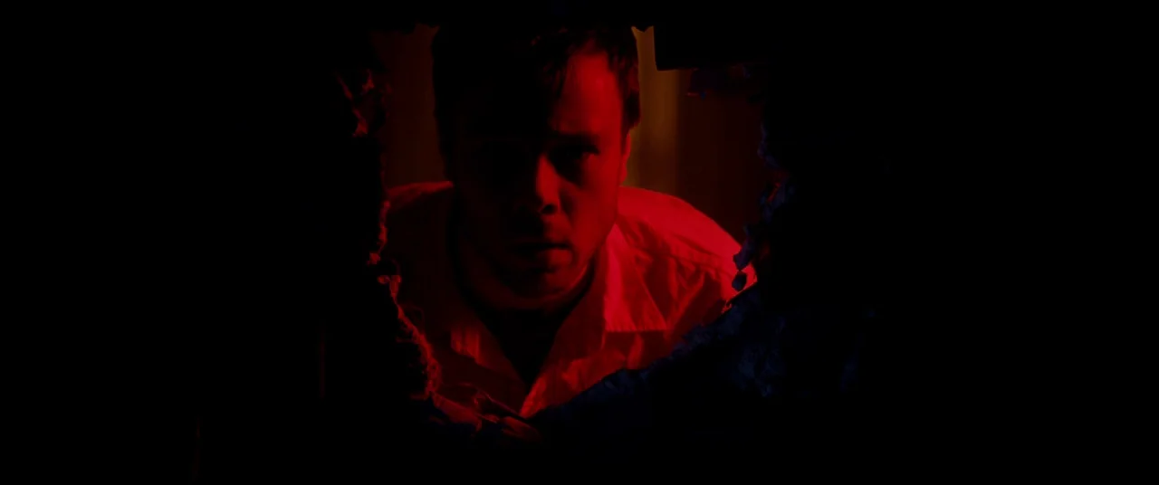 The Canal (2014) movie still: medium — A man looks through a jagged hole in a wall in a dark space. The subject is bathed in bright red light, while…