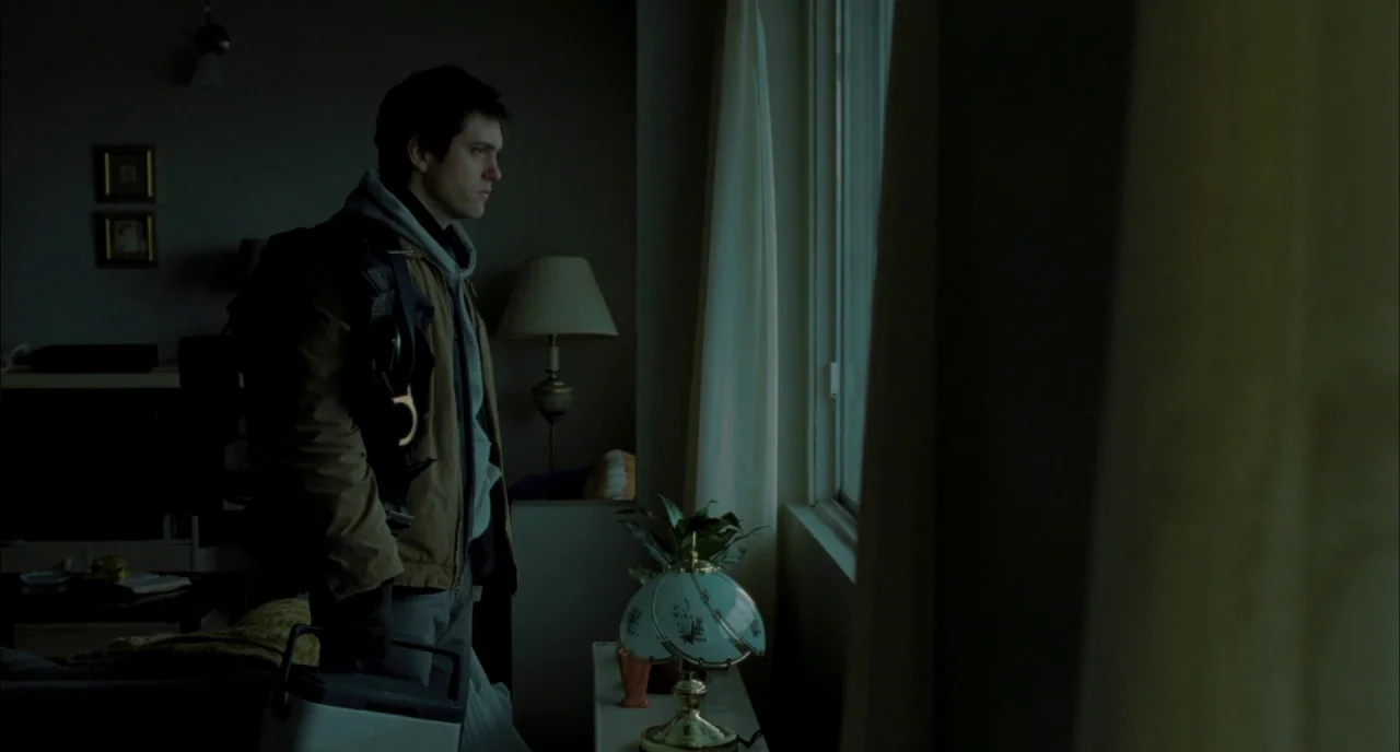 Incendies movie still: medium — A dimly lit room contains a table lamp, decorative plant, and framed wall art. A man wearing a backpack stands near…