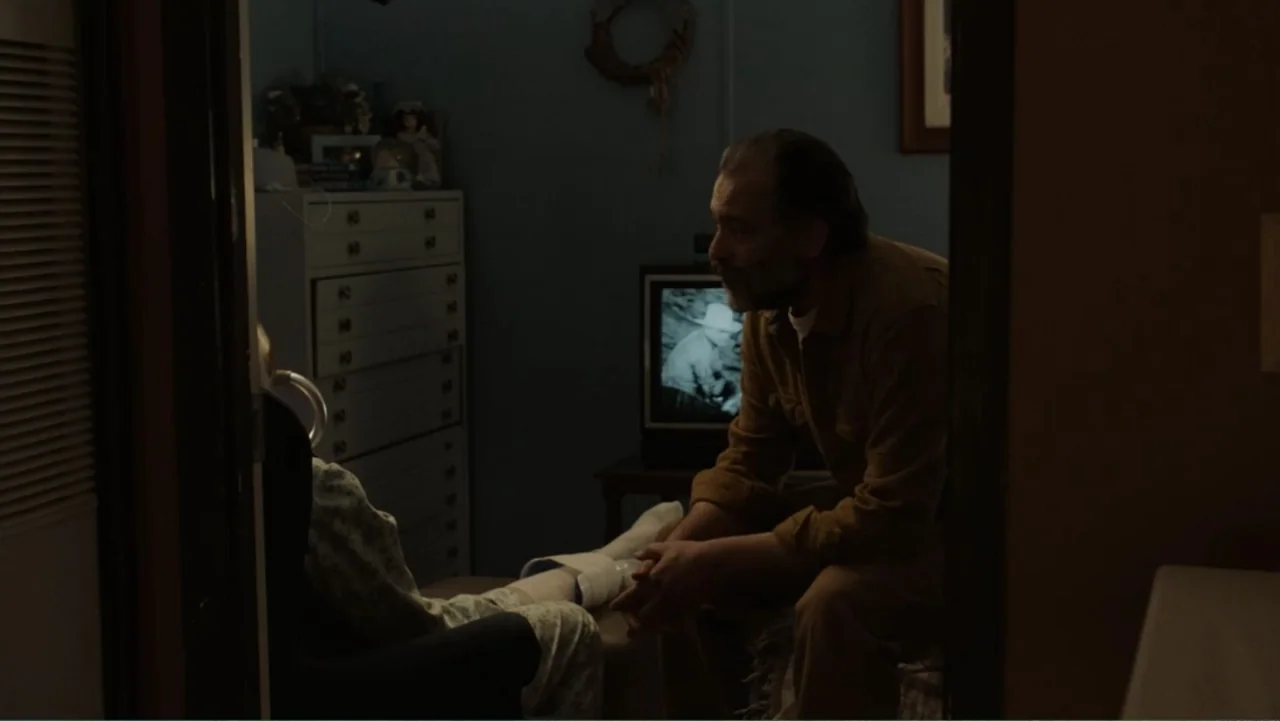 The Cleaner (2021) movie still: medium — A dimly lit bedroom features a tall chest of drawers and a glowing television. A man sits on a bed while…