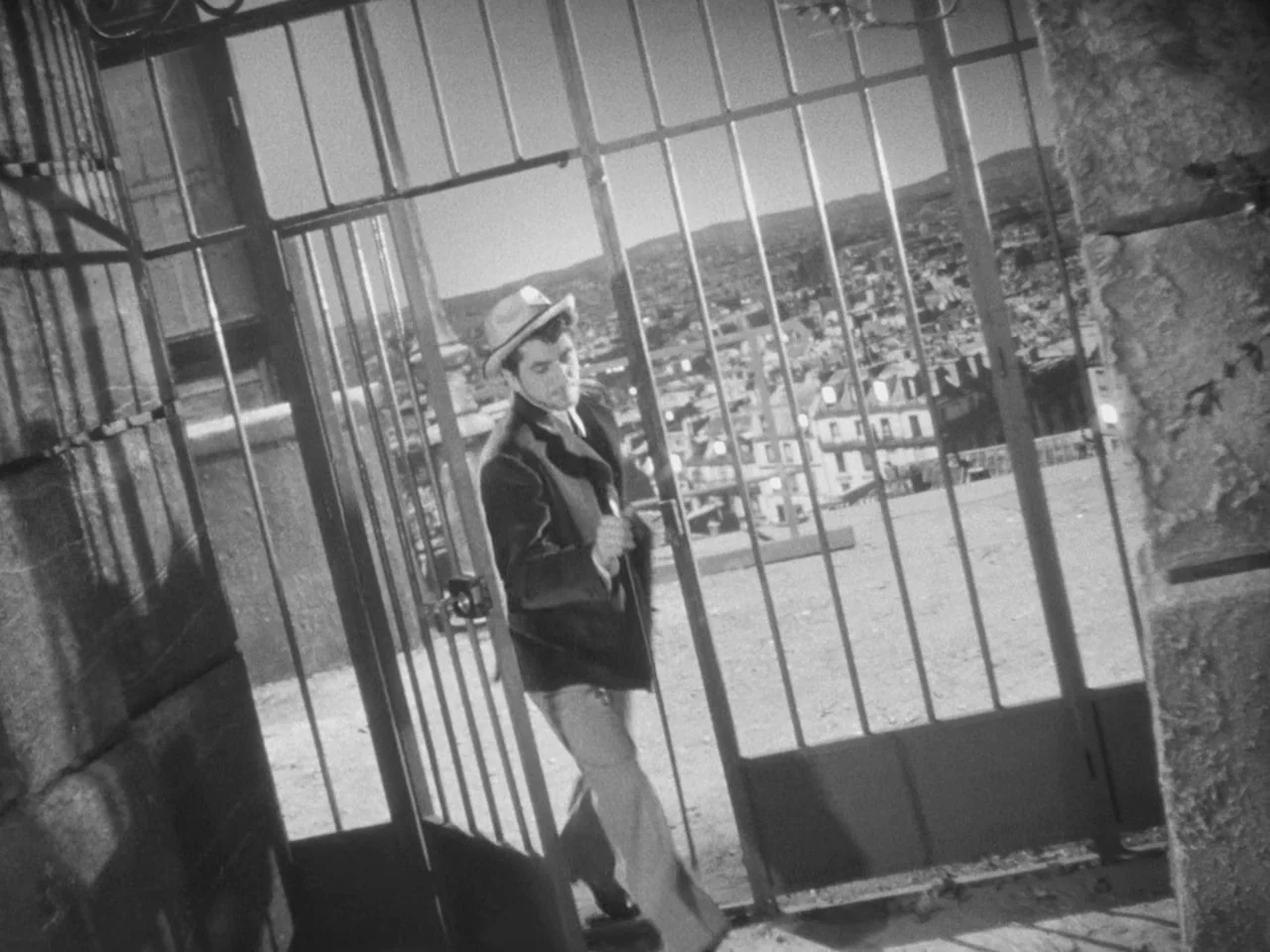 Le Plaisir (1952) movie still: fullbody — A metal gate stands open on a stone path overlooking a distant hillside city. A man in a blazer and flat…